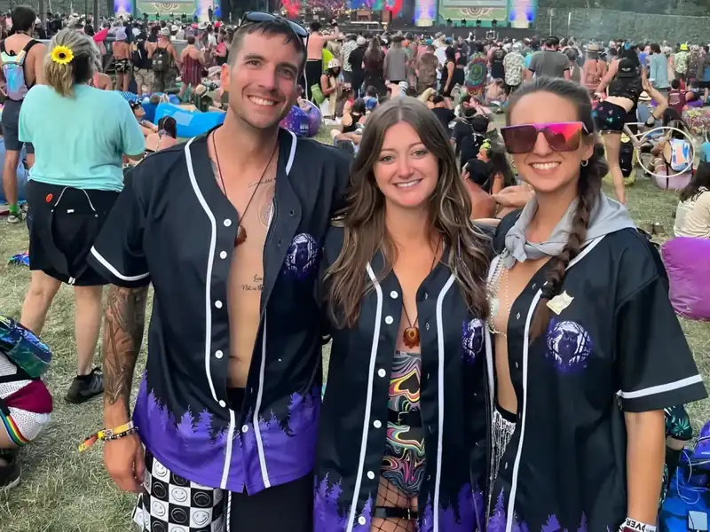 Three smiling customers wearing matching black baseball jerseys with purple forest artwork at a festival.